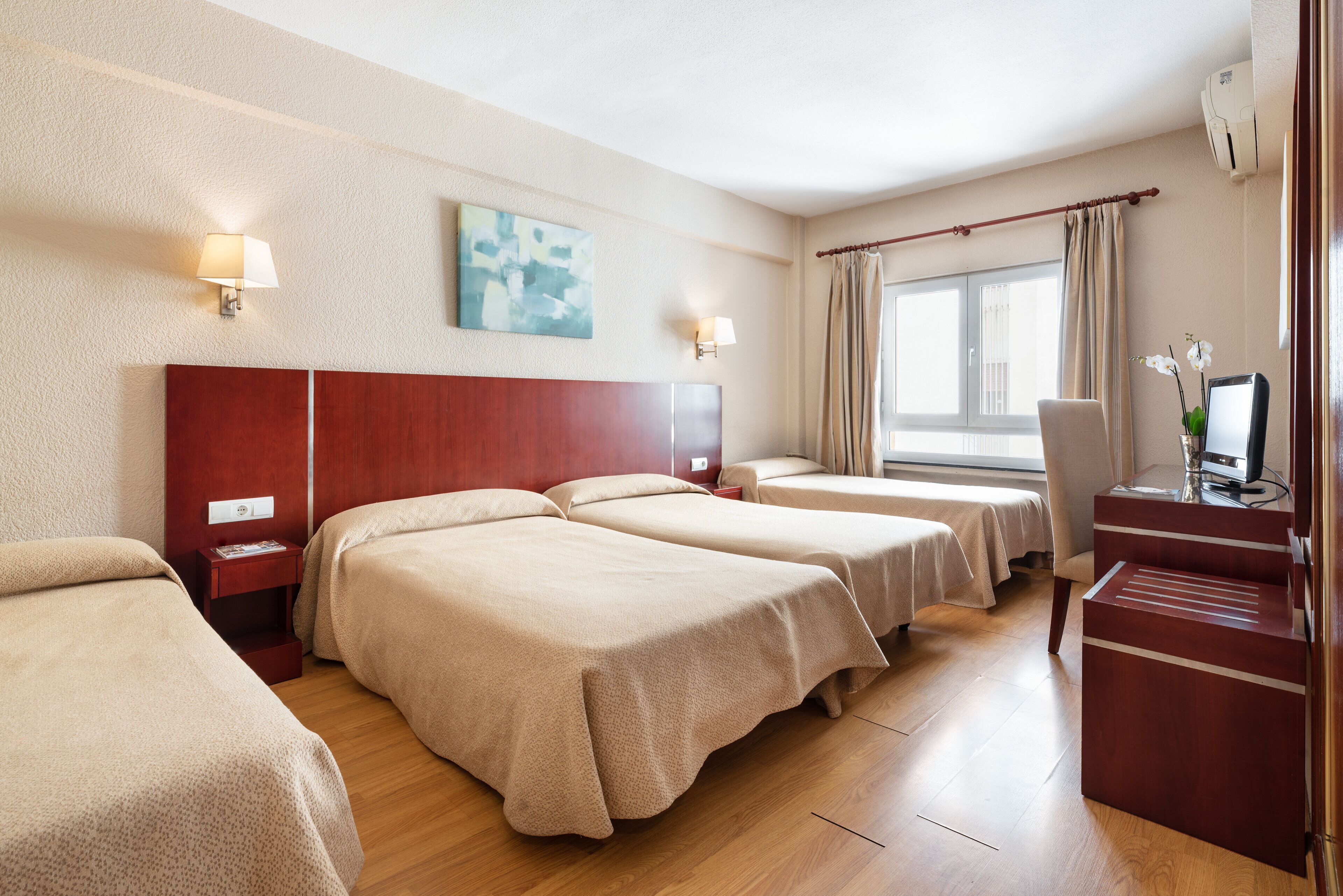 A hotel room with three beds, a desk with a chair, a window with curtains, and a painting on the wall.