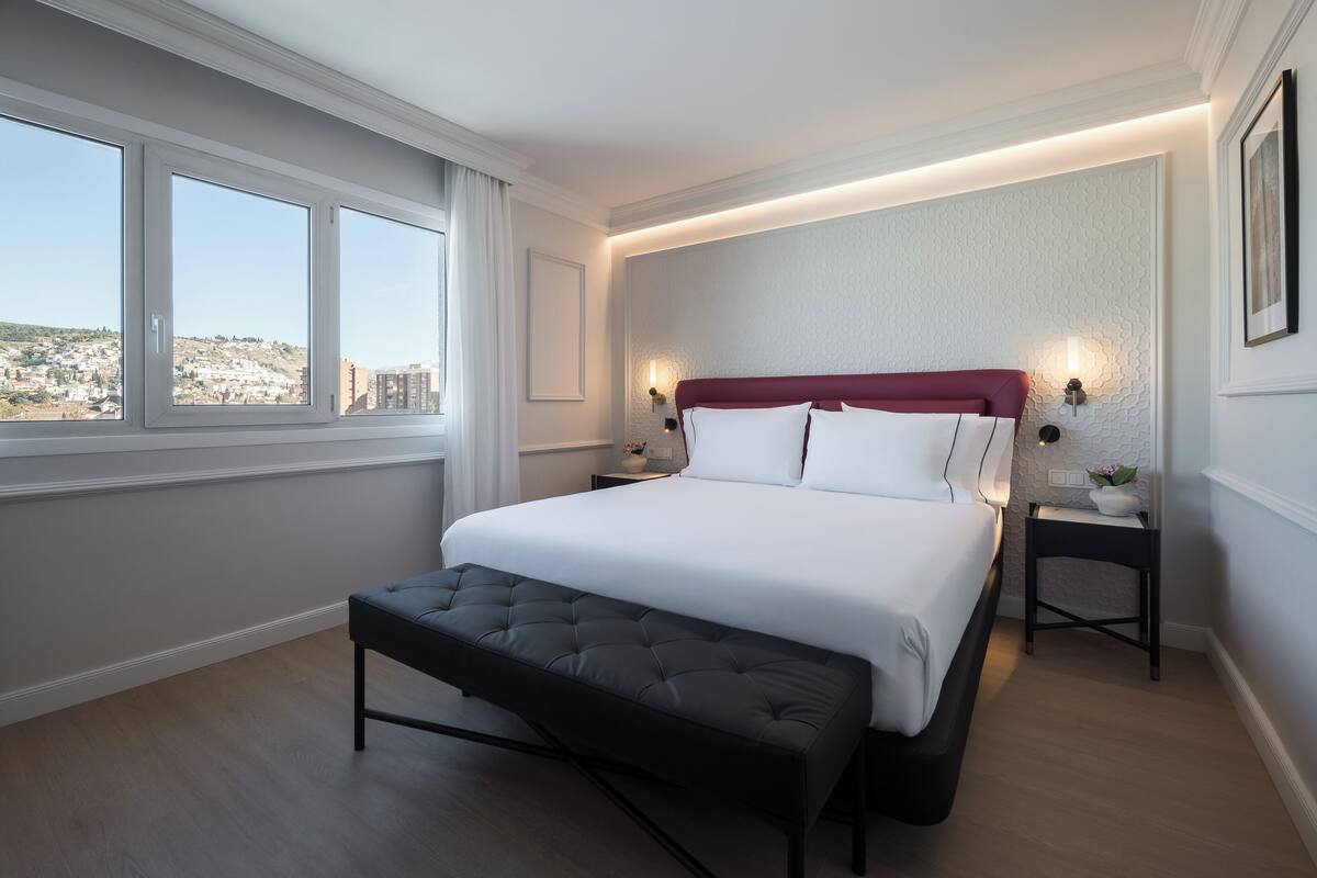 A modern hotel room with a large bed, a bench, a nightstand, and a view of the city.