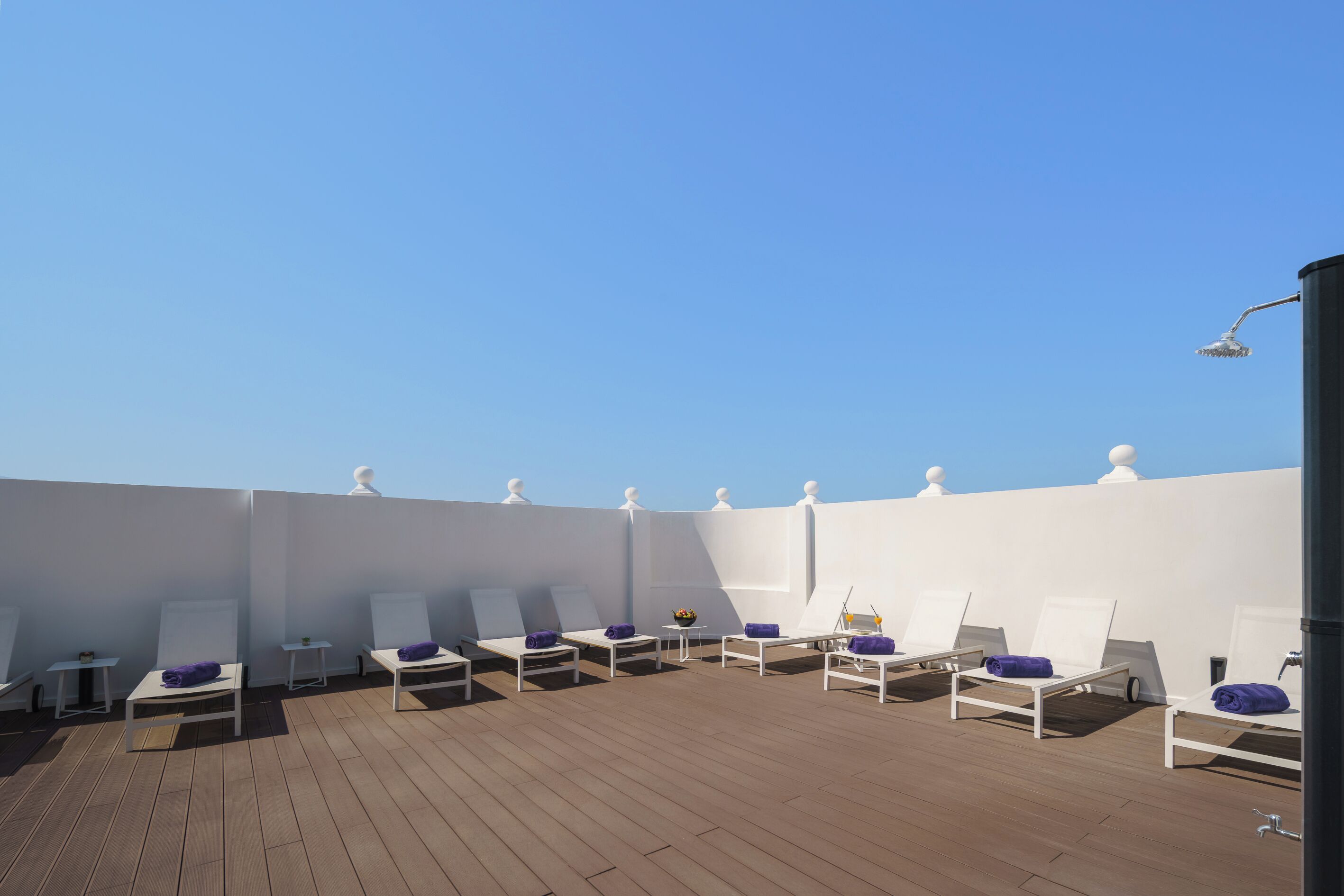 A rooftop lounge area with white lounge chairs and purple towels, a small table with a bowl of fruit, and a clear blue sky.