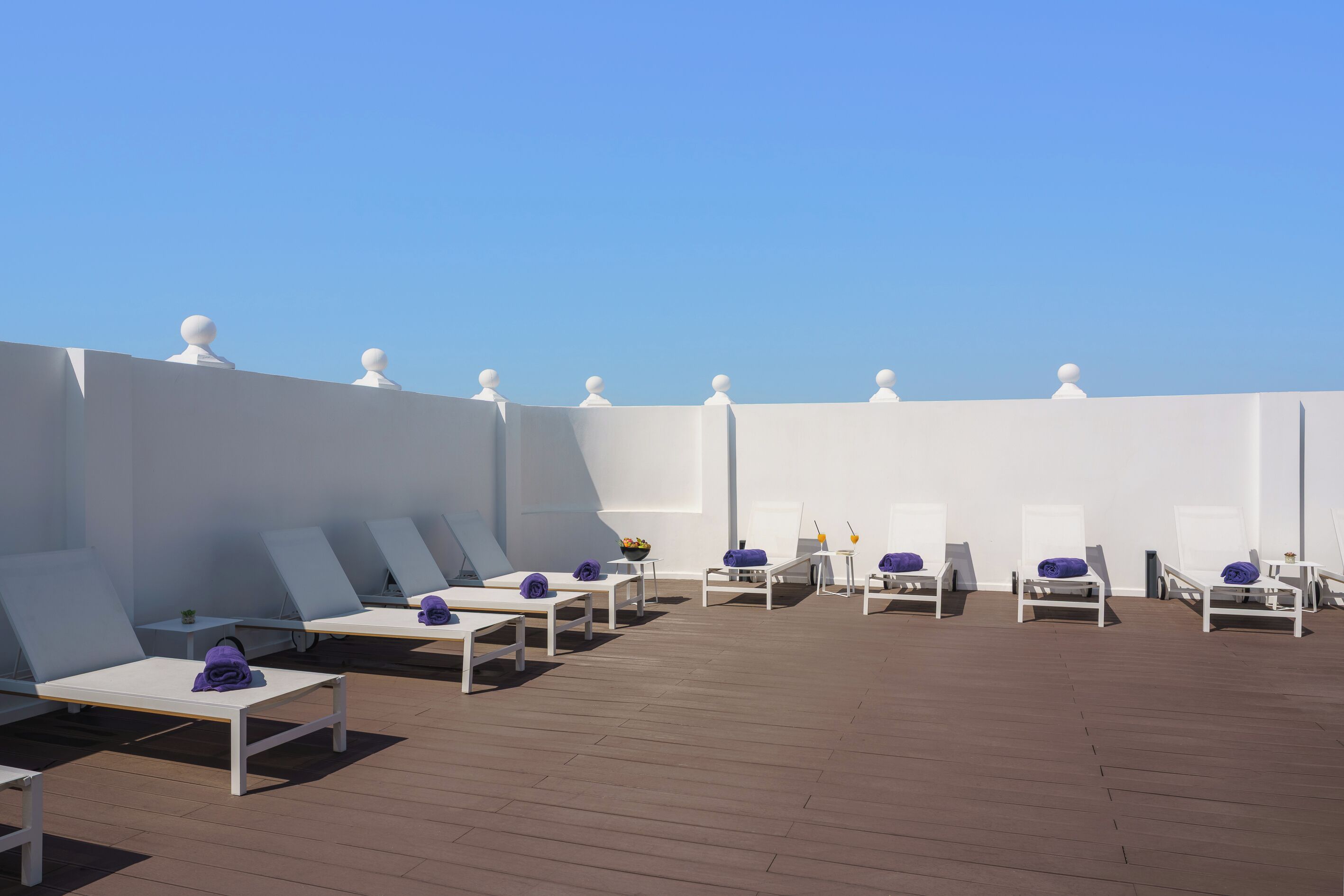 A rooftop lounge area with white lounge chairs and tables, arranged in rows, under a clear blue sky.