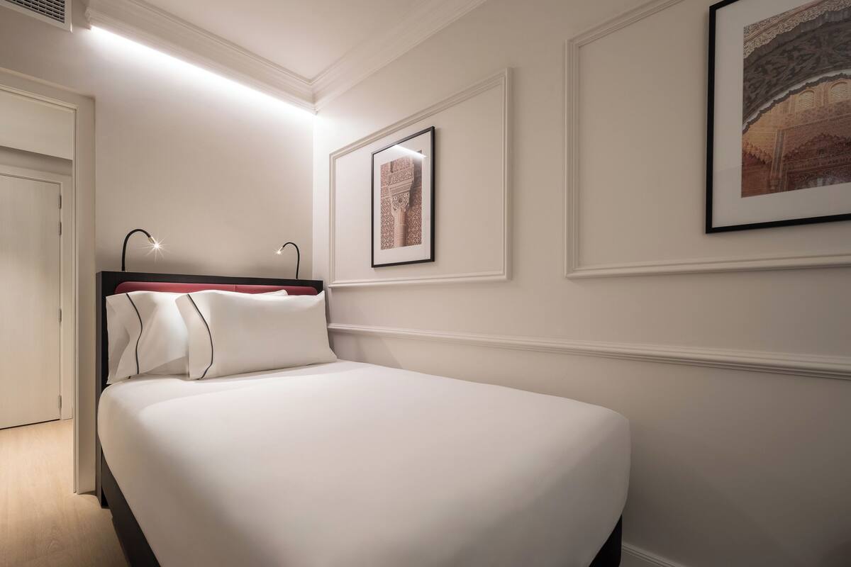 A neatly made bed in a hotel room with white walls, a framed picture on the wall, and a door to the left.