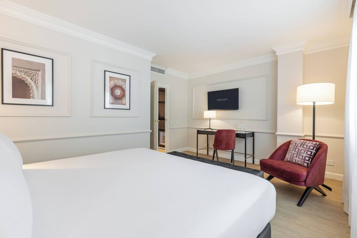 A hotel room with a large bed, a desk with a chair, a TV, and framed pictures on the wall.