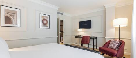 A hotel room with a large bed, a desk with a chair, a TV, and framed pictures on the wall.