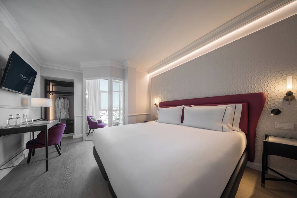 A modern hotel room with a large bed, a desk with a chair, a vanity with a mirror, and a view of the city.