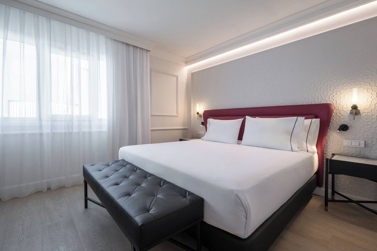 A modern hotel room with a large bed, a bench, a nightstand, and a wall-mounted lamp.