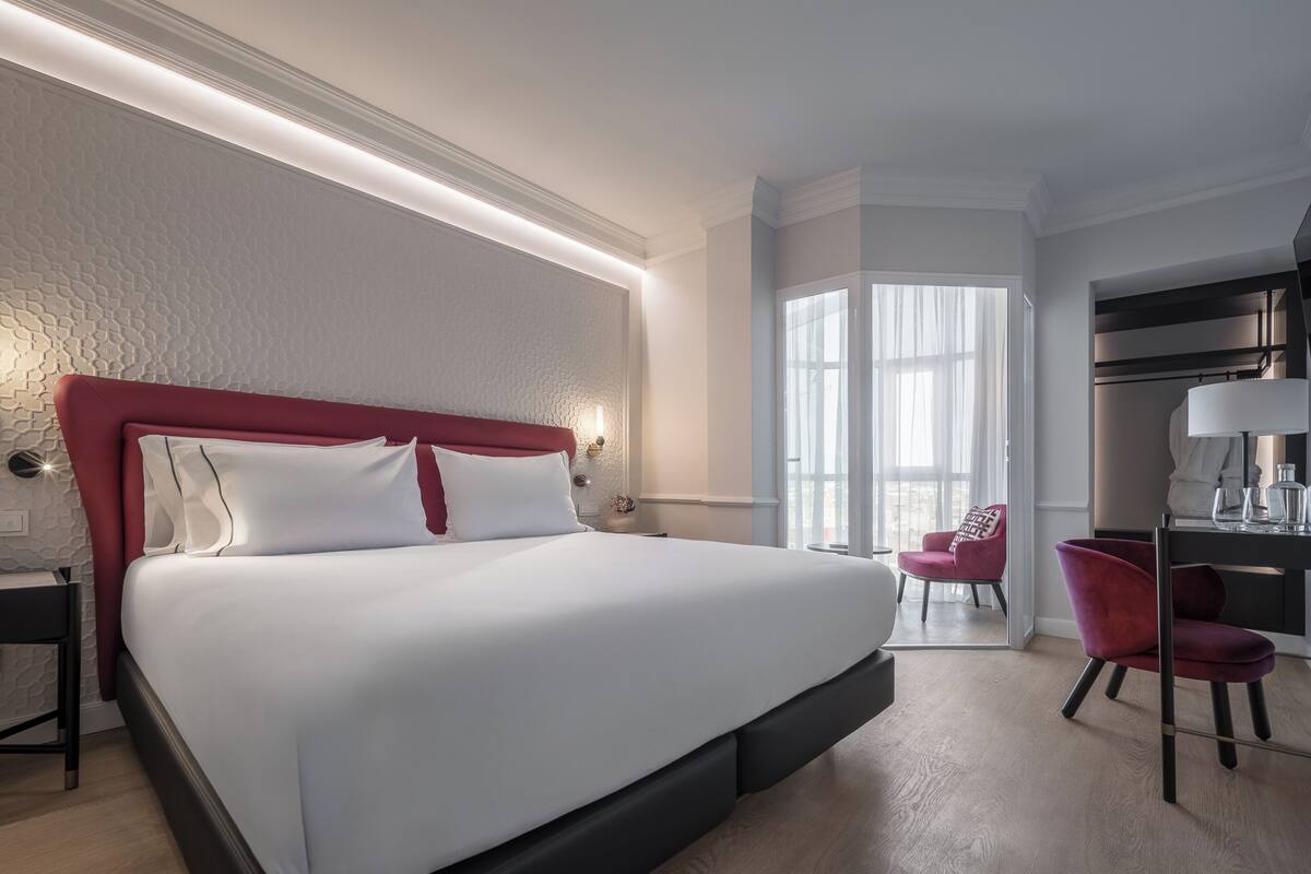 A modern hotel room with a large bed, a desk with a lamp, a chair, and a view of the city through large windows.