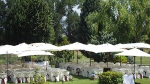 Filas de sillas blancas con cintas rojas, bajo sombrillas blancas, en un entorno al aire libre con árboles al fondo.
