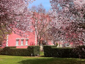 Un edificio rojo con detalles blancos y fachada simétrica, rodeado de setos cuidados y cerezos en flor.