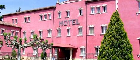 A pink building with a 'HOTEL' sign, multiple windows, and a well-maintained lawn with a tall coniferous tree.