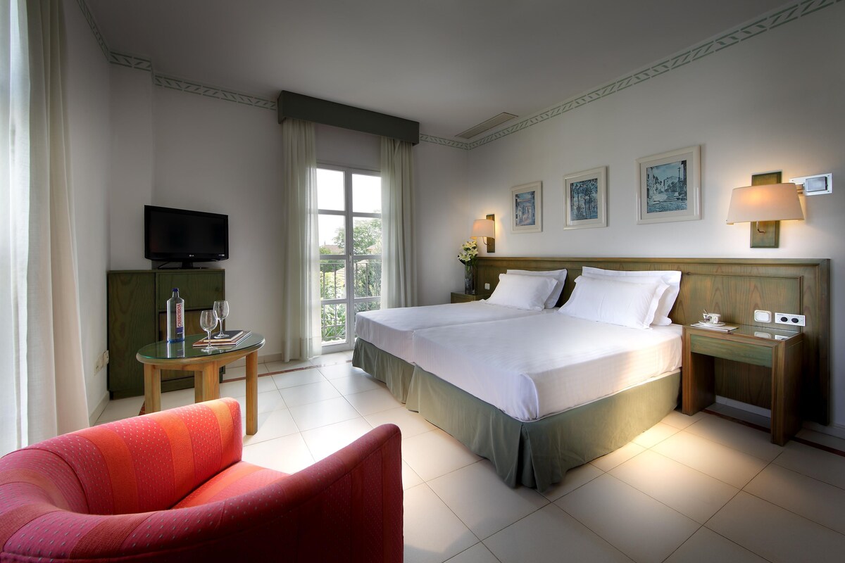 Habitación de hotel con una cama grande, un televisor sobre una mesita, una silla y una ventana con cortinas.