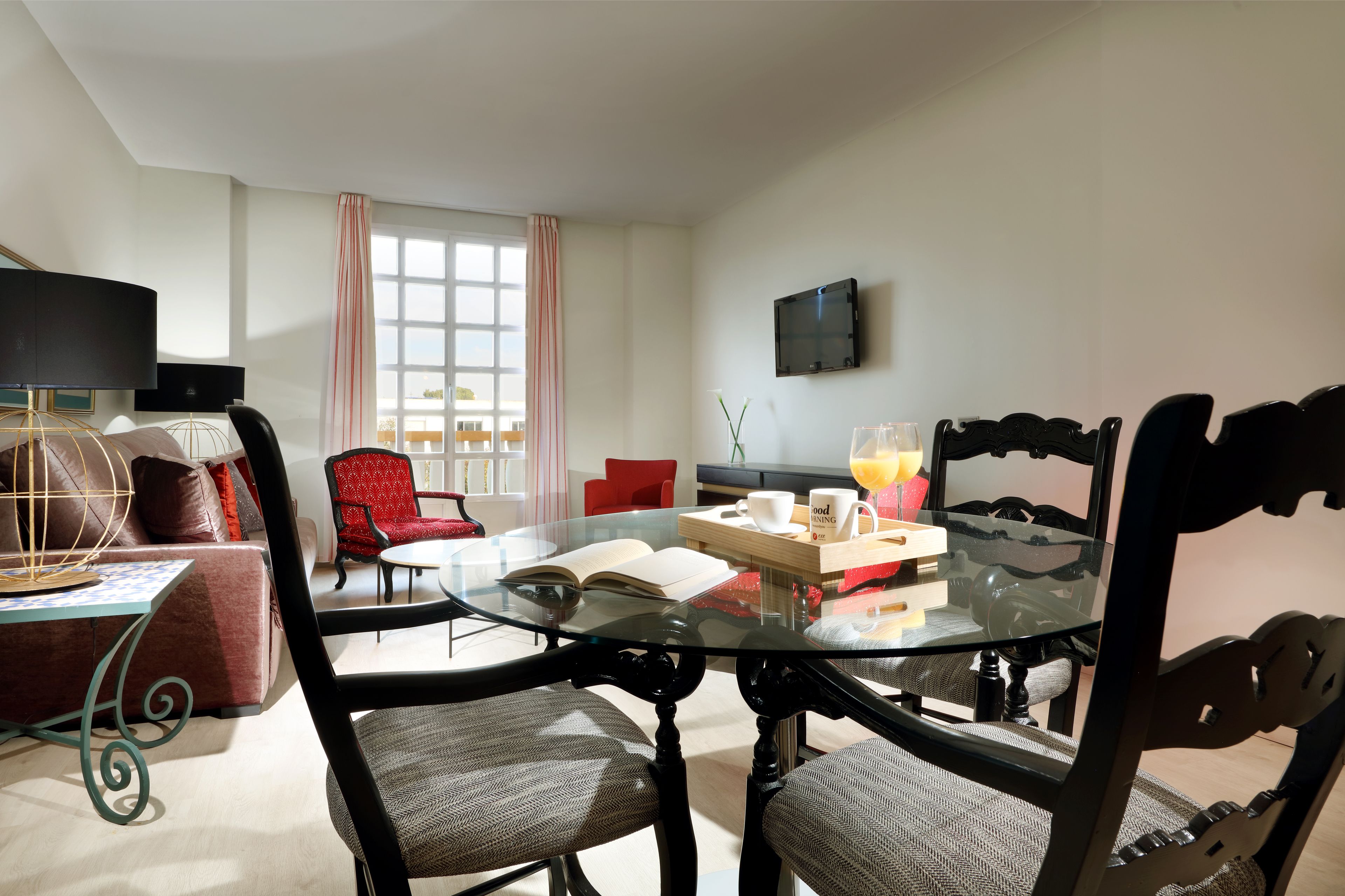 A modern living room with a glass table, red and black chairs, a sofa, and a TV on the wall.