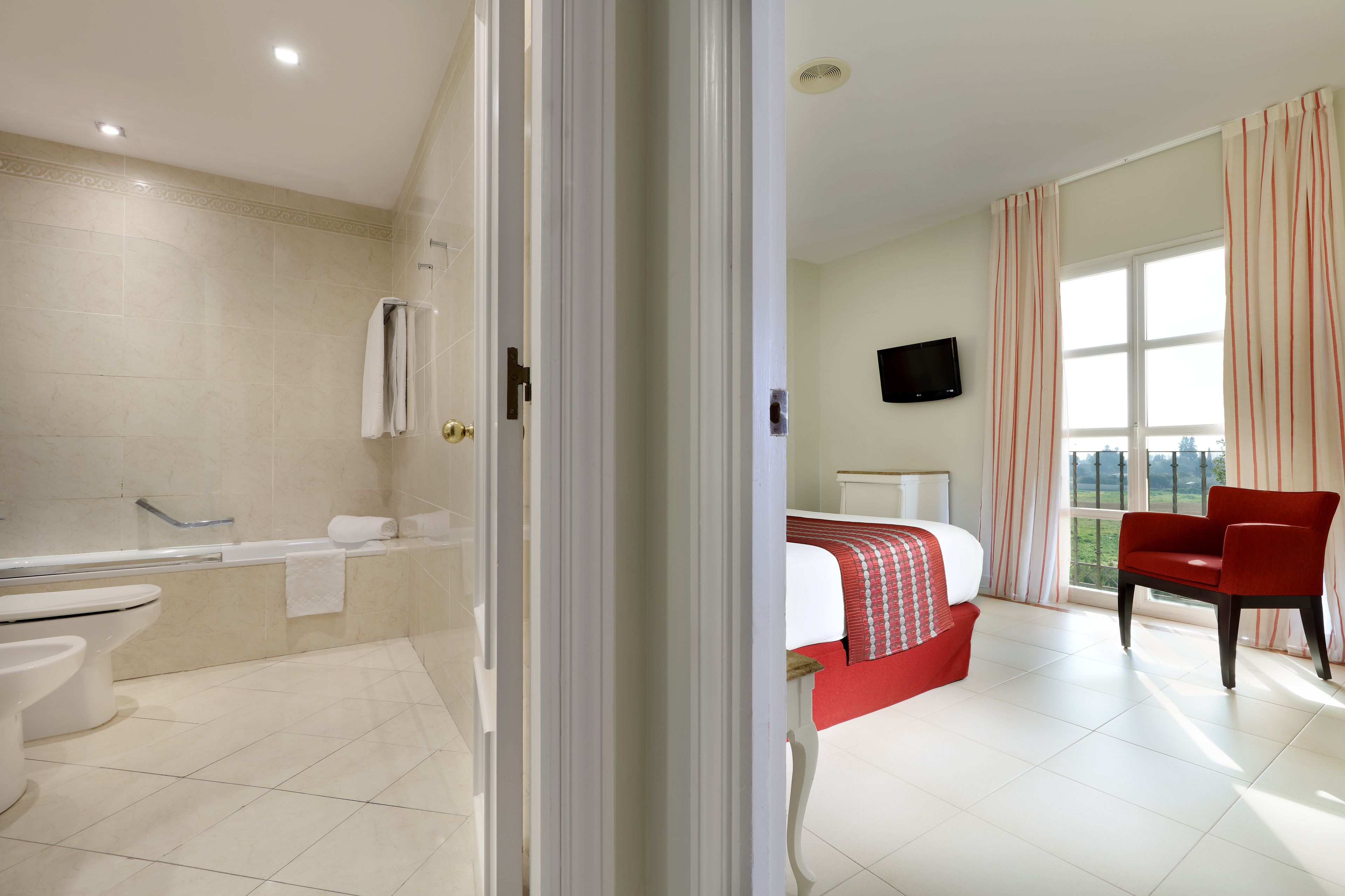 A hotel room with a bathroom, a bed, a red chair, and a TV.