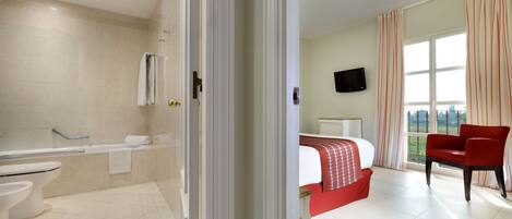 A hotel room with a bathroom, a bed, a red chair, and a TV.