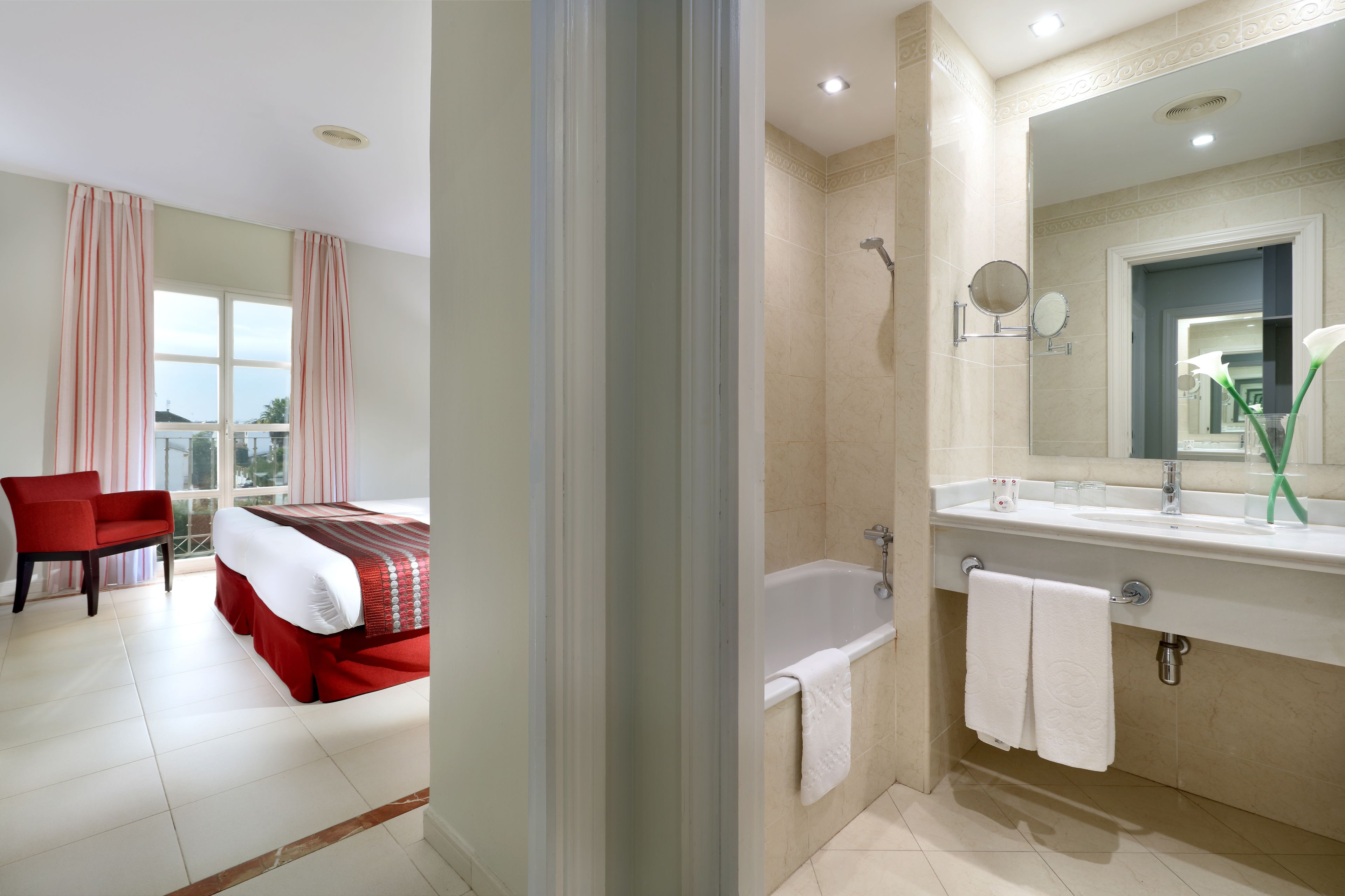 Una habitación de hotel con cama, una silla roja, baño con bañera y un espejo grande.