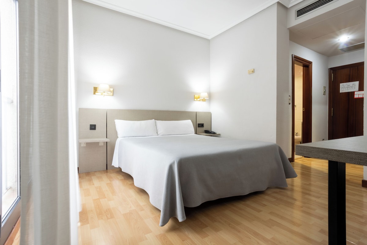A hotel room with a bed, bedside tables, and wall-mounted lamps.