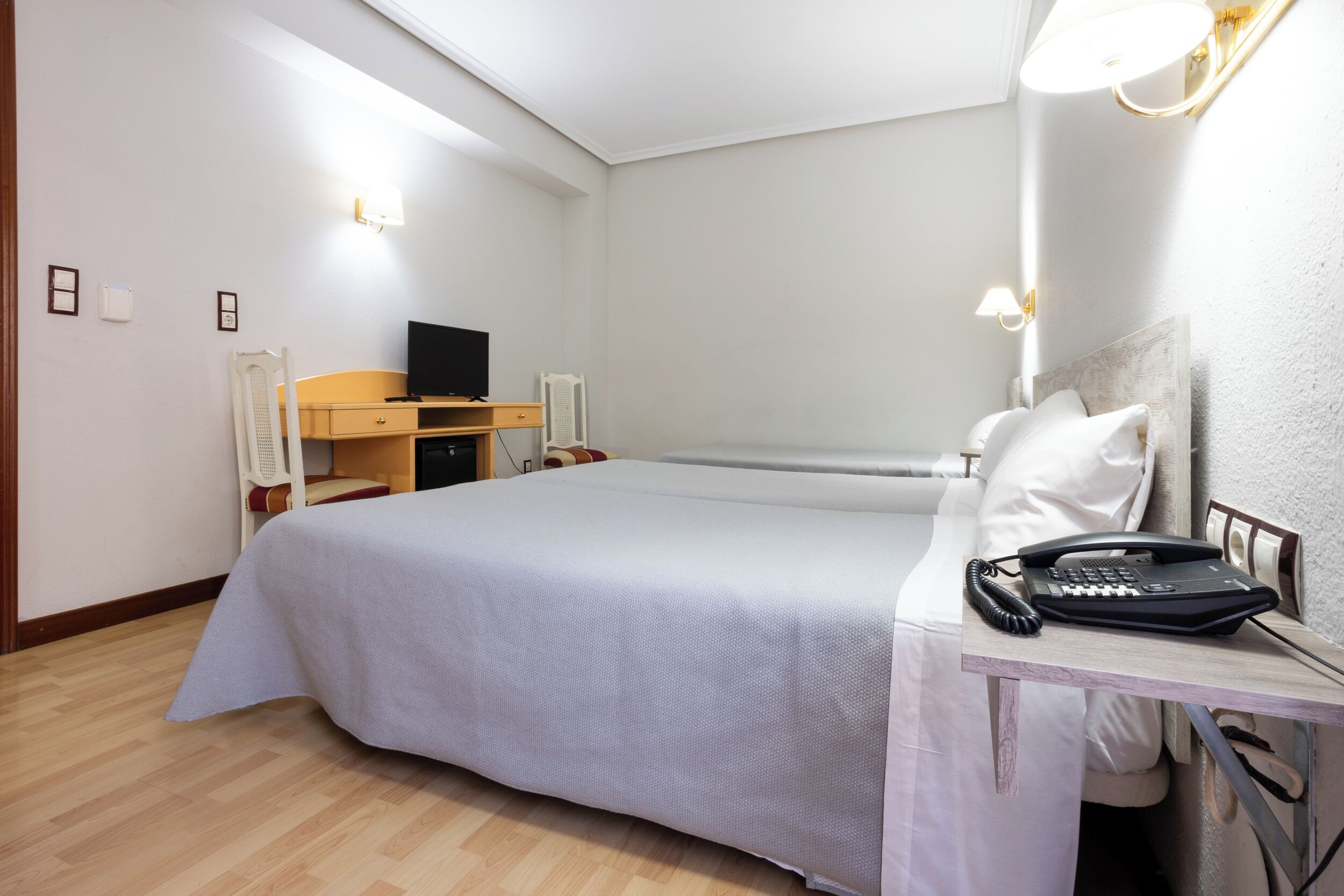 A hotel room with a bed, bedside table, lamp, and a desk with a telephone.