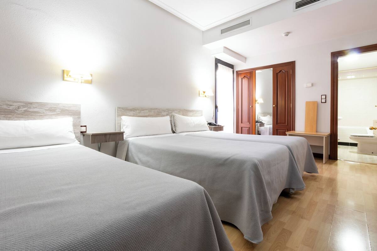 A hotel room with two beds, a wooden floor, a mirror, a bedside table, and a door leading to a bathroom.