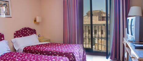 A hotel room with two beds, a TV, a desk, and a balcony with a view of buildings.