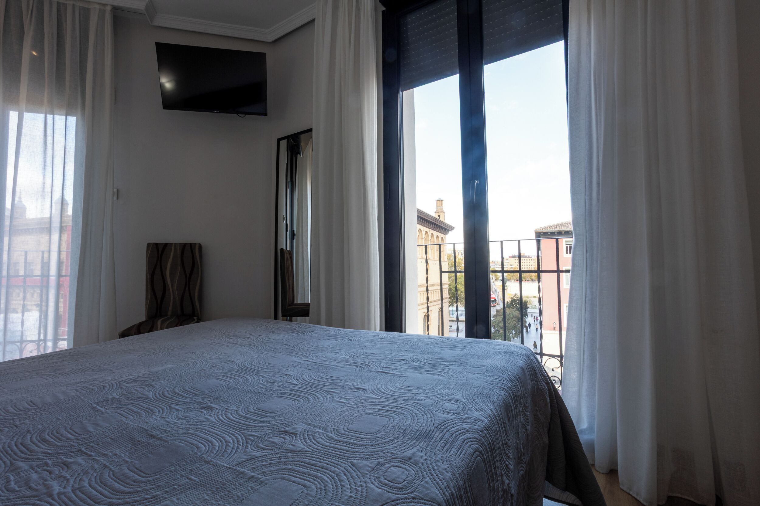 Een slaapkamer met een groot bed, een wandtelevisie en een balkon met gordijnen.