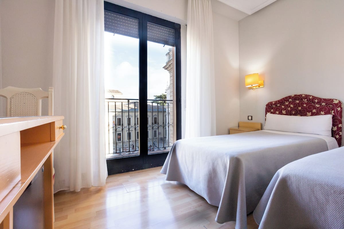A hotel room with two beds, a wooden dresser, a nightstand, a wall-mounted lamp, and a balcony with a view of buildings.