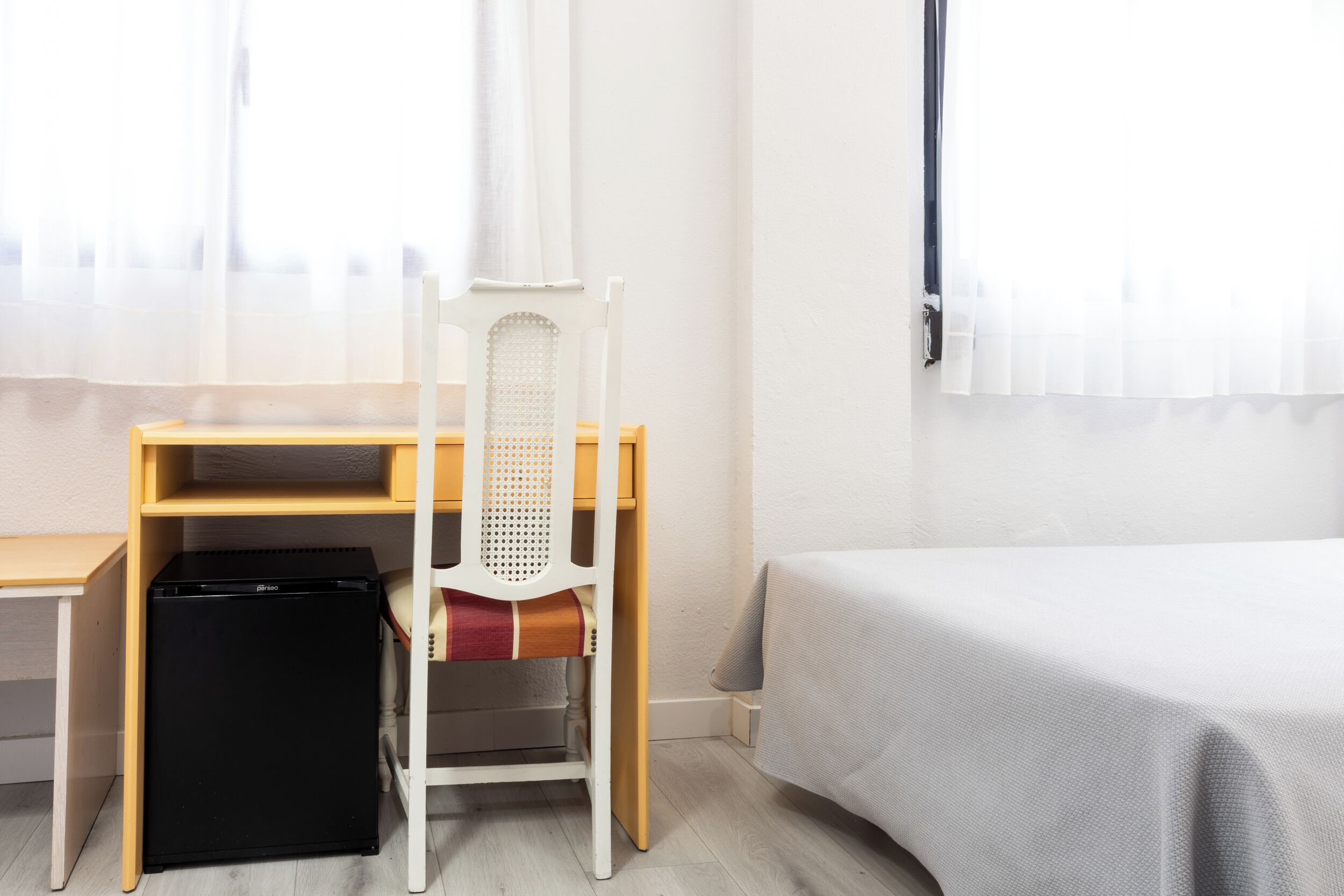 A bedroom with a bed, a desk with a chair, a small black refrigerator, and a window with sheer curtains.