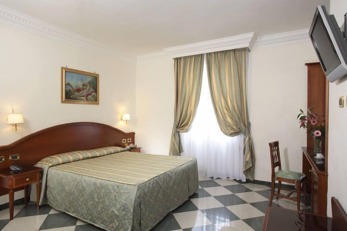 A hotel room with a large bed, two bedside tables, a TV mounted on the wall, and a window with curtains.