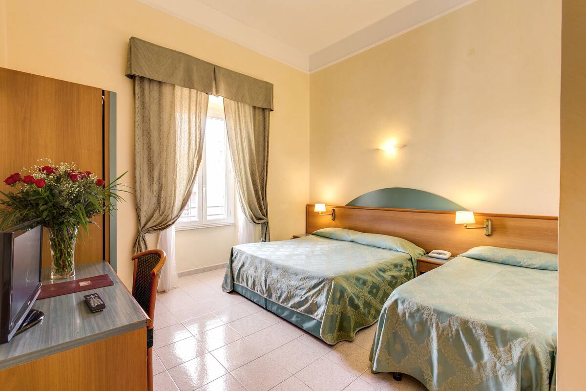 A hotel room with two beds, a desk with a TV and remote, a vase with flowers, and a window with curtains.