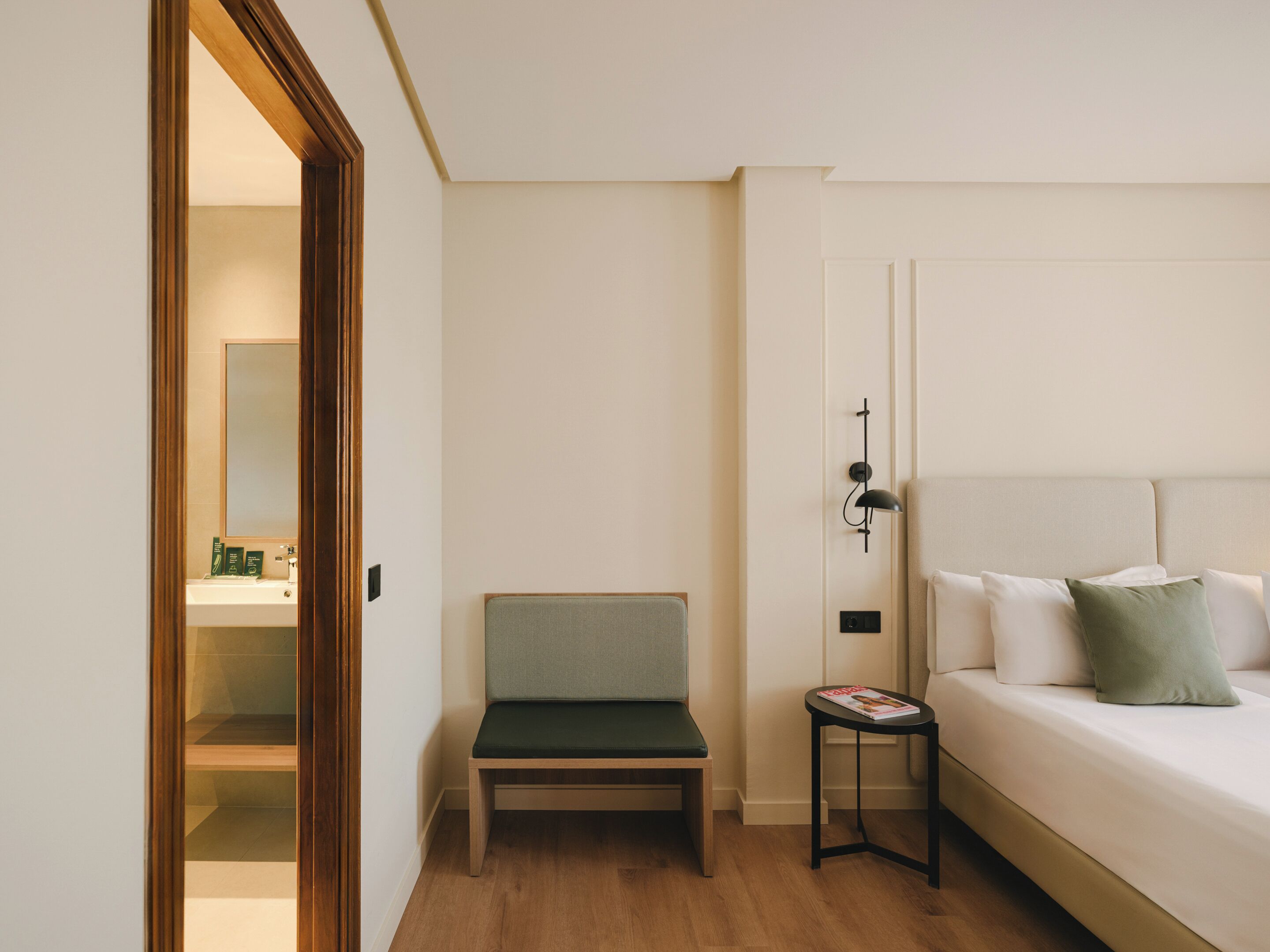 A hotel room with a bed, a bedside table, a chair, a small table, and a mirror.