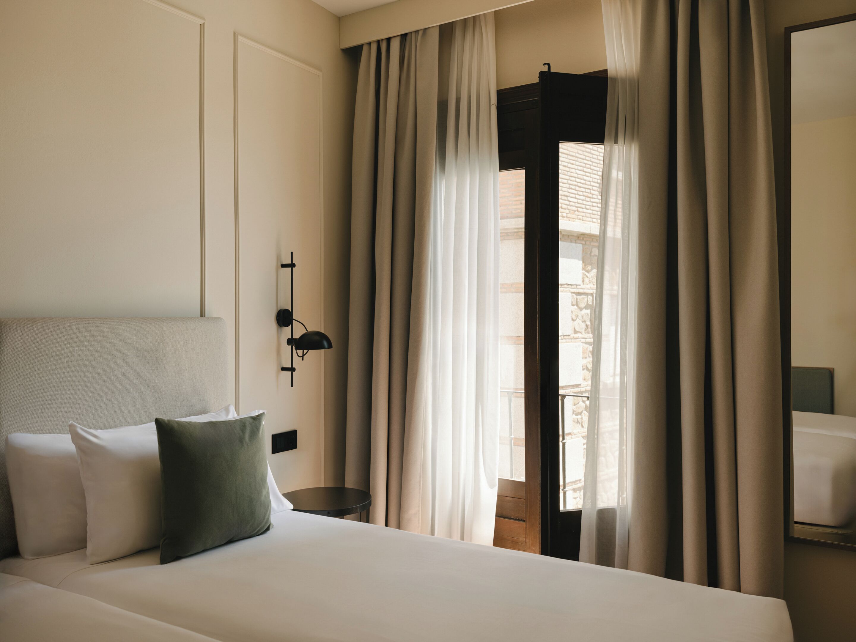 A hotel room with a bed, a bedside table, a lamp, and a window with curtains.