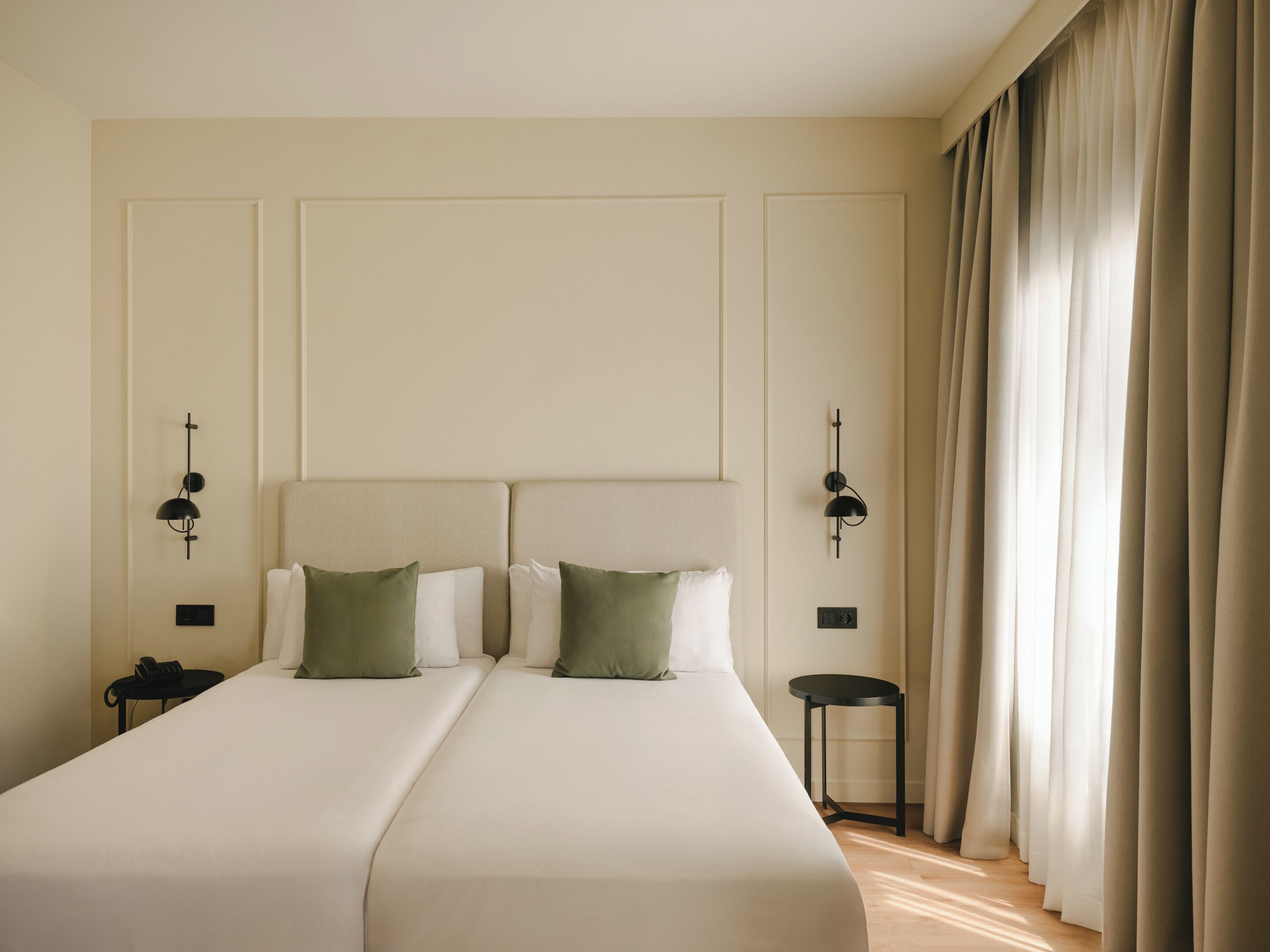 Una cama doble con dos almohadas, una mesita de noche, una lámpara de pared y una ventana con cortinas.
