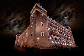 Um edifício grande e histórico com vários andares e uma torre imponente, iluminado à noite.