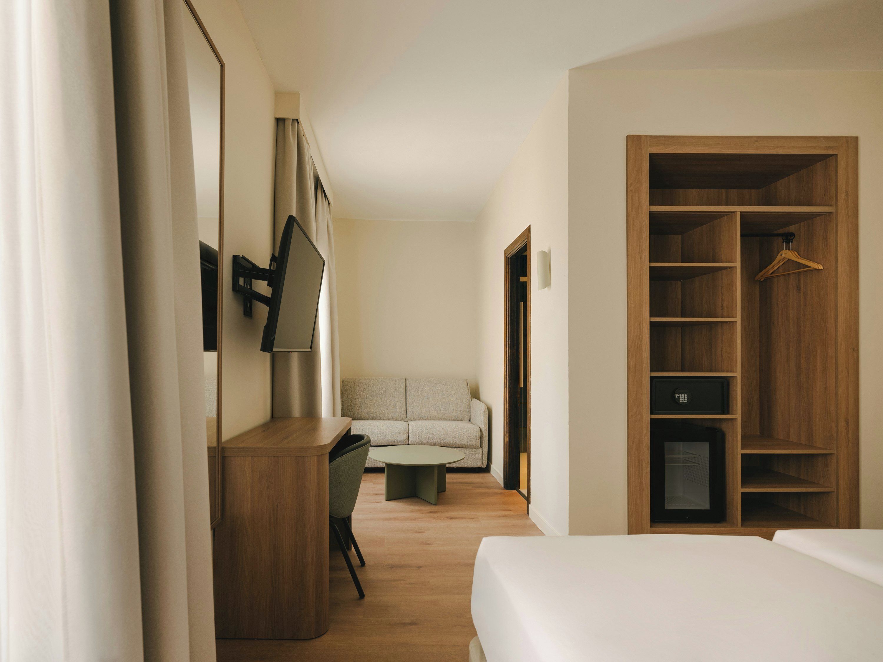 Una habitación de hotel con cama, escritorio, silla, sofá, armario y televisor montado en la pared.