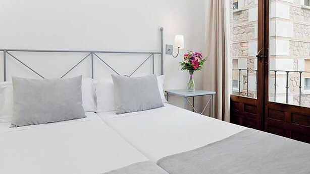Ein Hotelzimmer mit einem Bett, einem Nachttisch, einer Vase mit Blumen und Blick auf das Gebäude draußen.