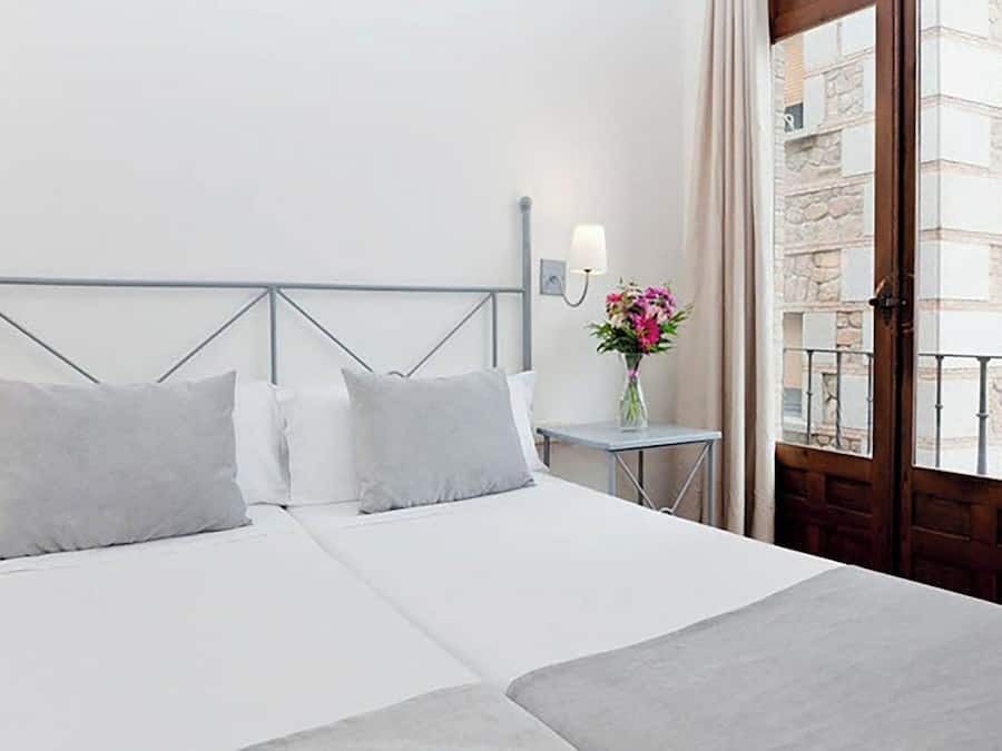 Habitación de hotel con cama, mesita de noche, un florero con flores y vistas del edificio exterior.