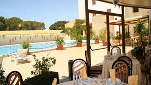 Un área de comedor al aire libre con mesas dispuestas para dos, con vista a una piscina, plantas en macetas y un edificio con techo de tejas.