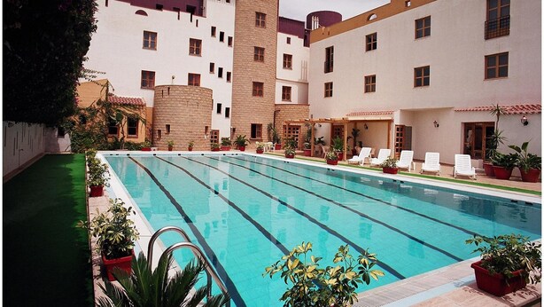 Una piscina con una estructura que recuerda a un castillo al fondo, rodeada de plantas en macetas y sillas de descanso.