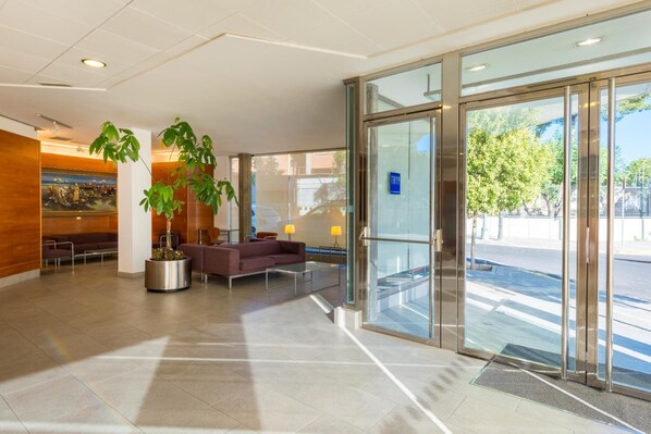 A modern lobby with a reception desk, seating area, and large glass doors leading outside. - Hotel Port Feria Valencia (Valencia)