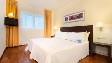 A hotel room with a large bed, a flat-screen TV mounted on the wall, a painting above the bed, and a desk with a lamp.