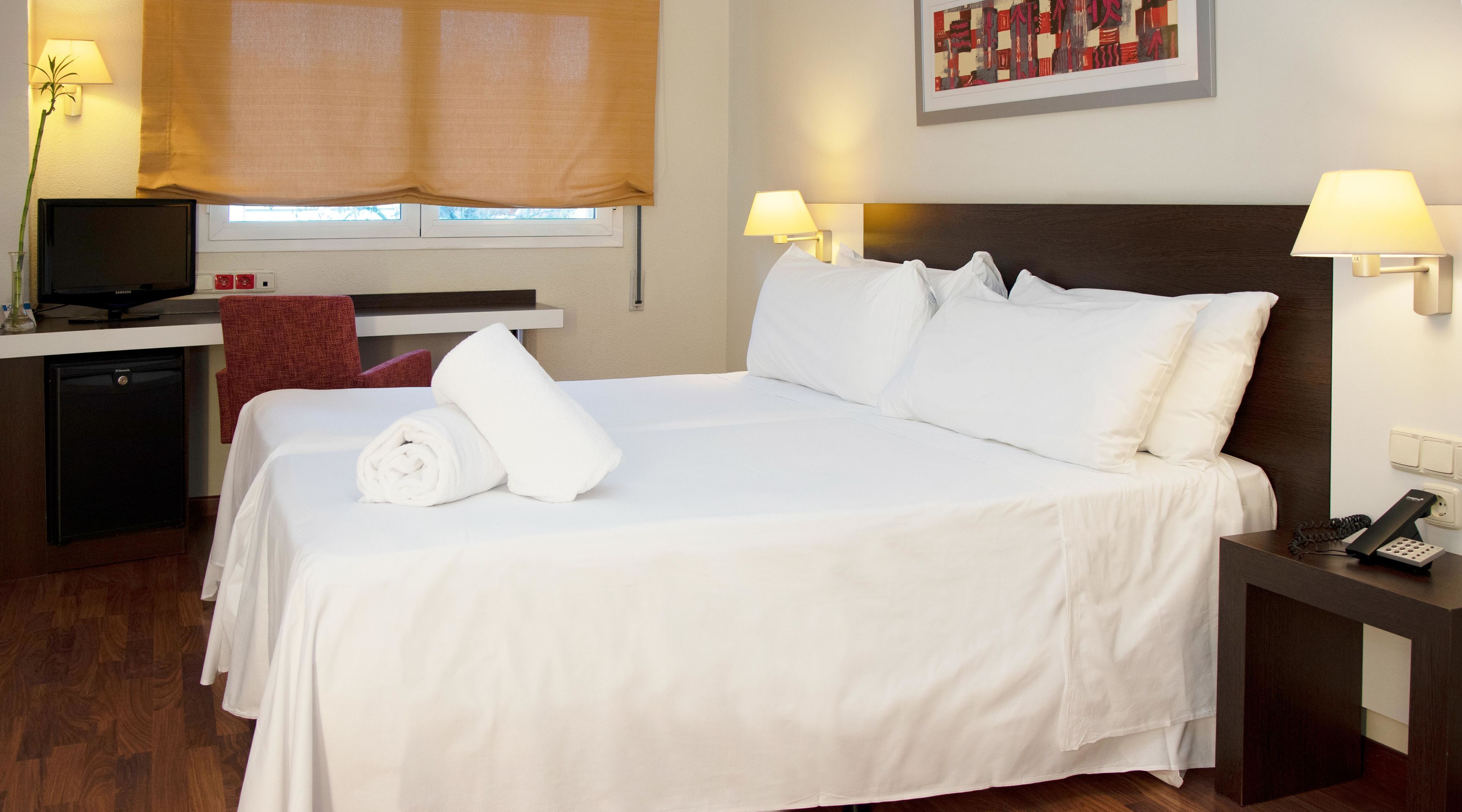 A neatly made bed with white linens, a bedside table with a lamp, a small desk with a computer, and a window with a blind.