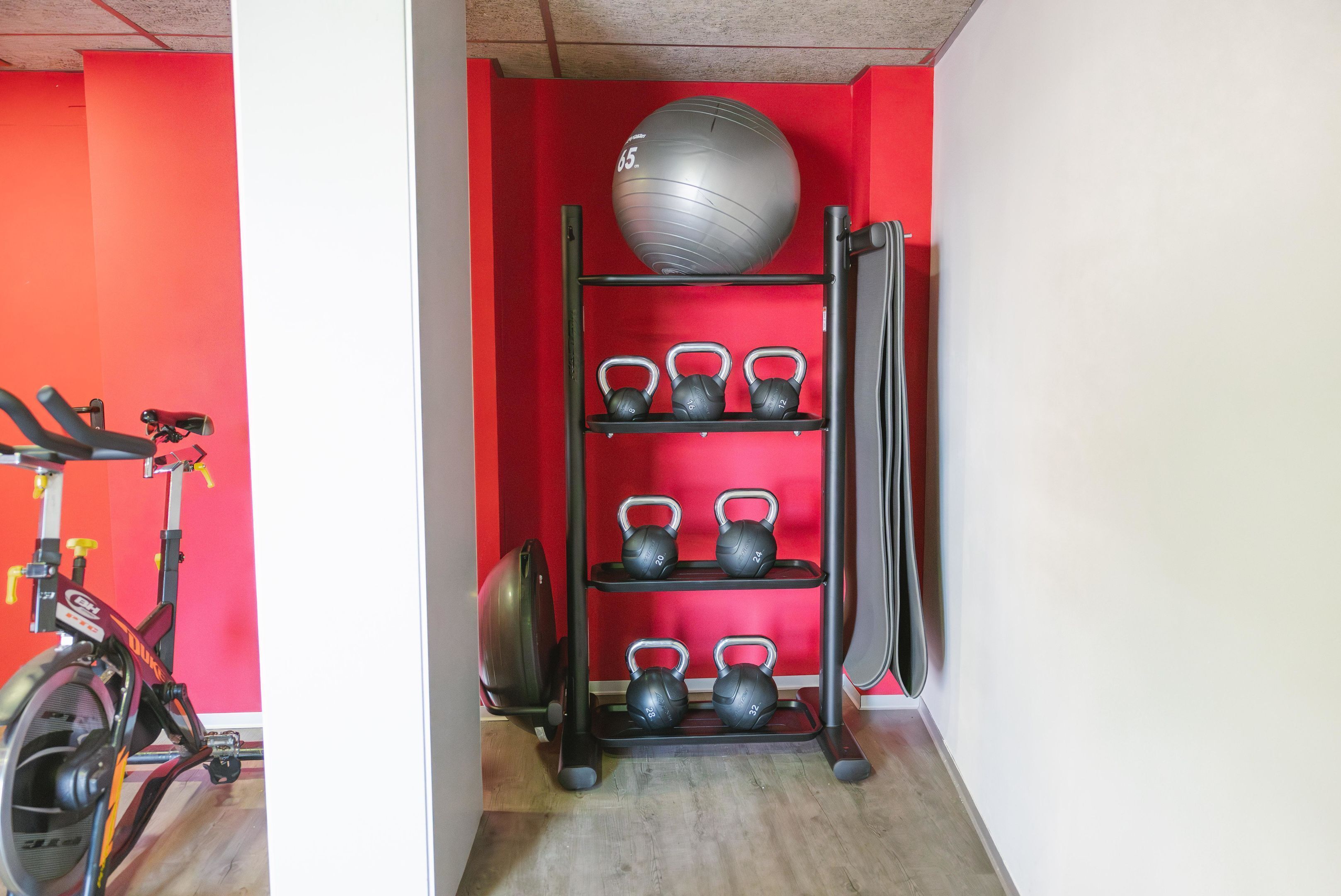 Un gimnasio con una pared roja, una bicicleta estática, un soporte para kettlebells y una pelota de ejercicios grande.