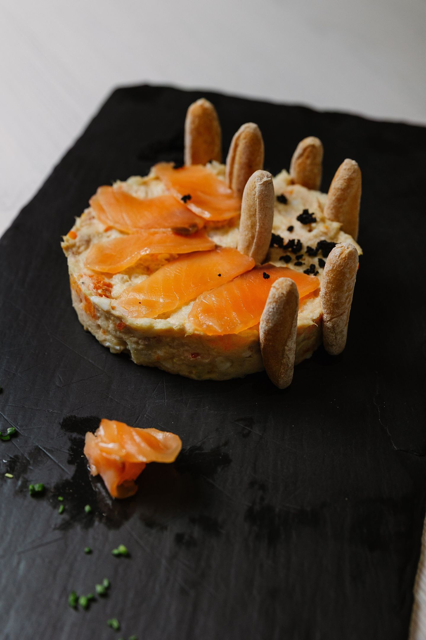 Una tabla de pizarra negra con un sándwich redondo cubierto de salmón ahumado y galletas.