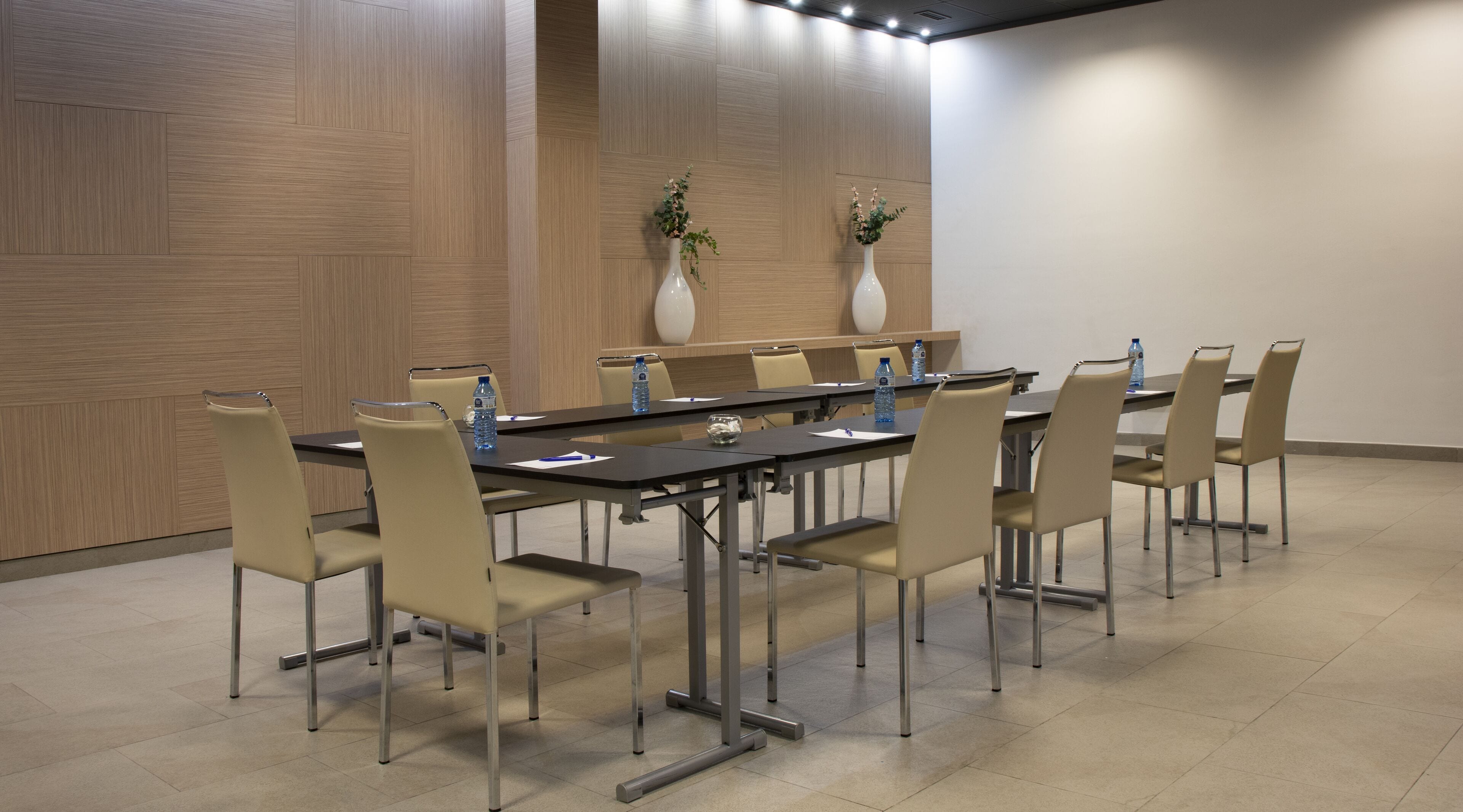 Un salón de conferencias con una larga mesa, sillas color beige y dos jarrones blancos con plantas.