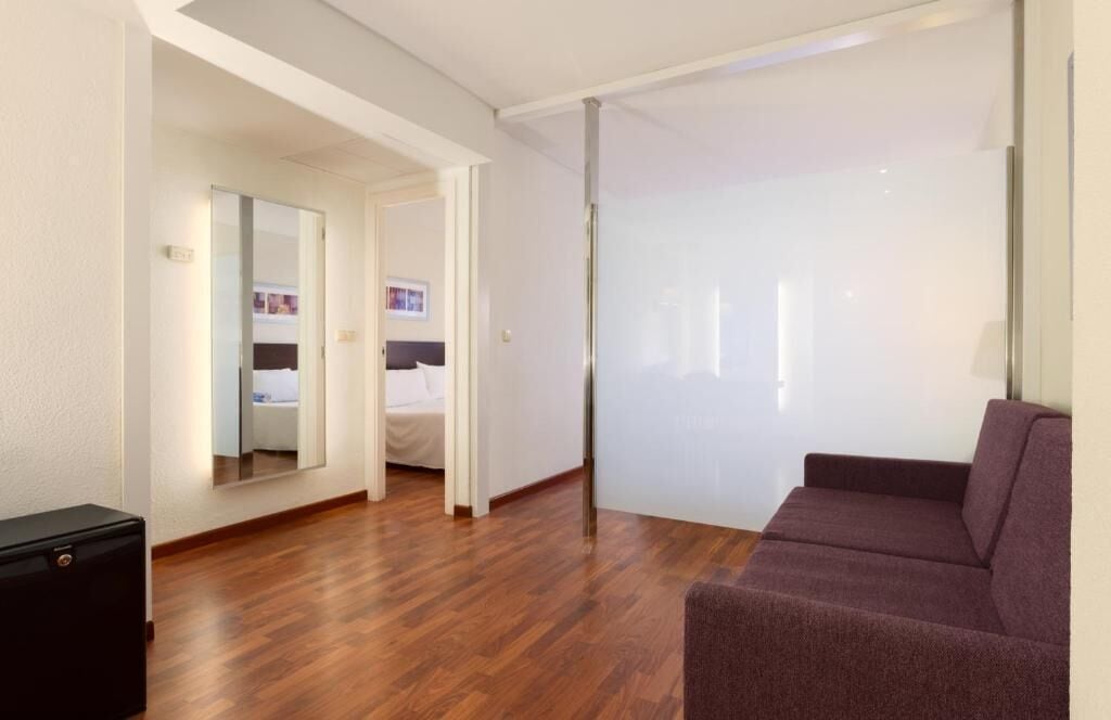 A modern hotel room with a wooden floor, a purple sofa, a bed, a large mirror, and a sliding door.