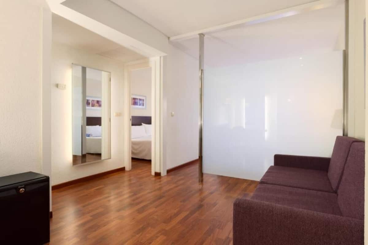 Una habitación de hotel moderna con piso de madera, un sofá púrpura, una cama, un espejo grande y una puerta corredera.