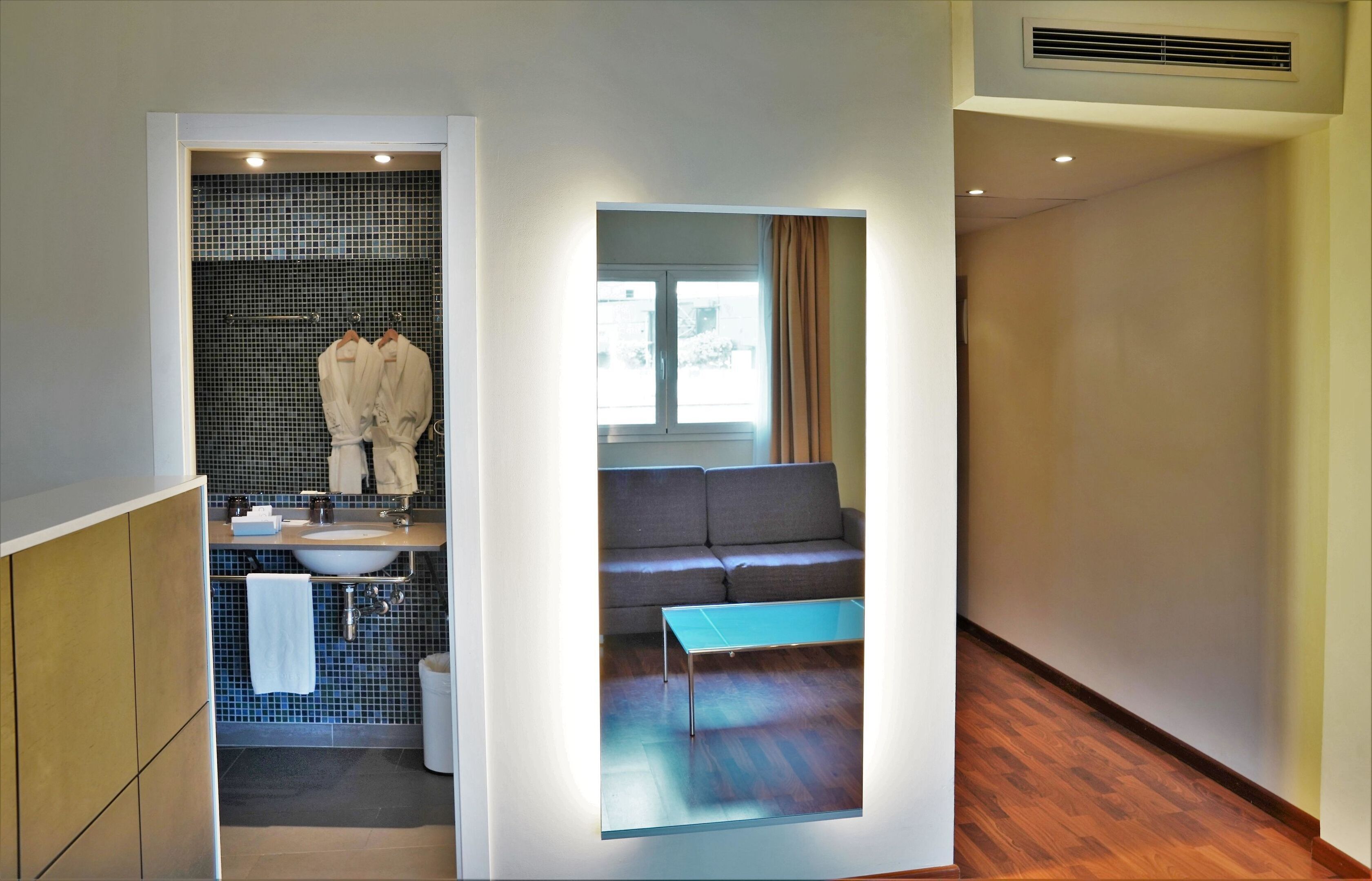 Een moderne badkamer met een wastafel, spiegel en handdoeken. Grenzend is een zithoek met een bank en een raam.