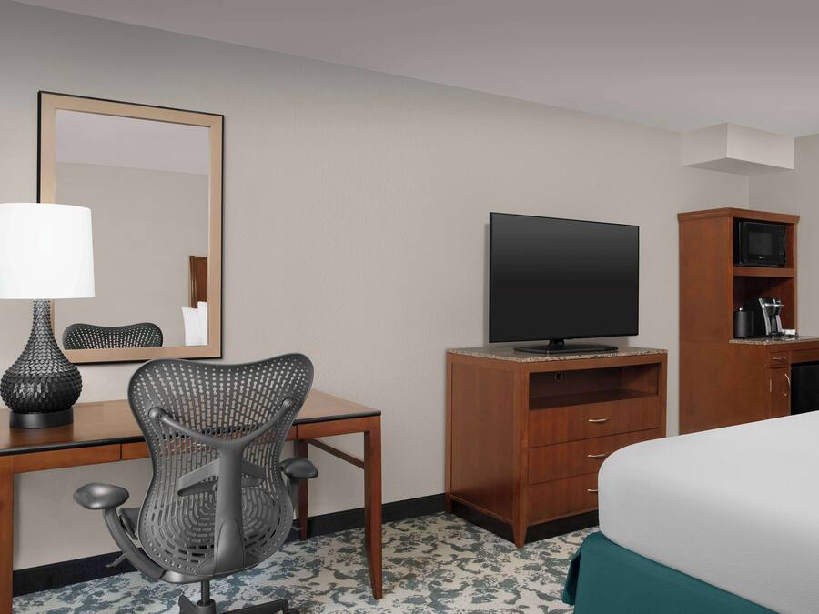 Quarto de hotel com cama, escrivaninha com cadeira, televisão, guarda-roupa e espelho.
