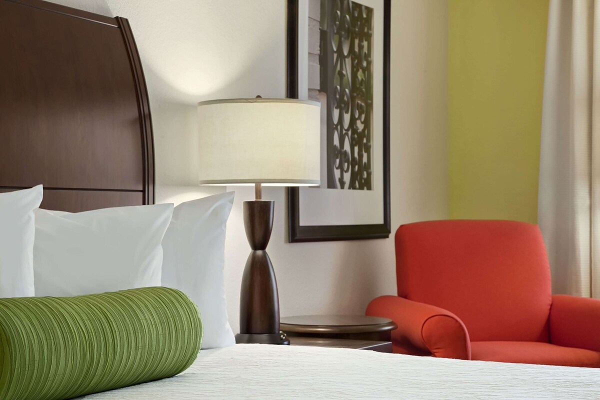 A hotel room with a bed, pillows, a lamp, a red chair, and a framed artwork on the wall.