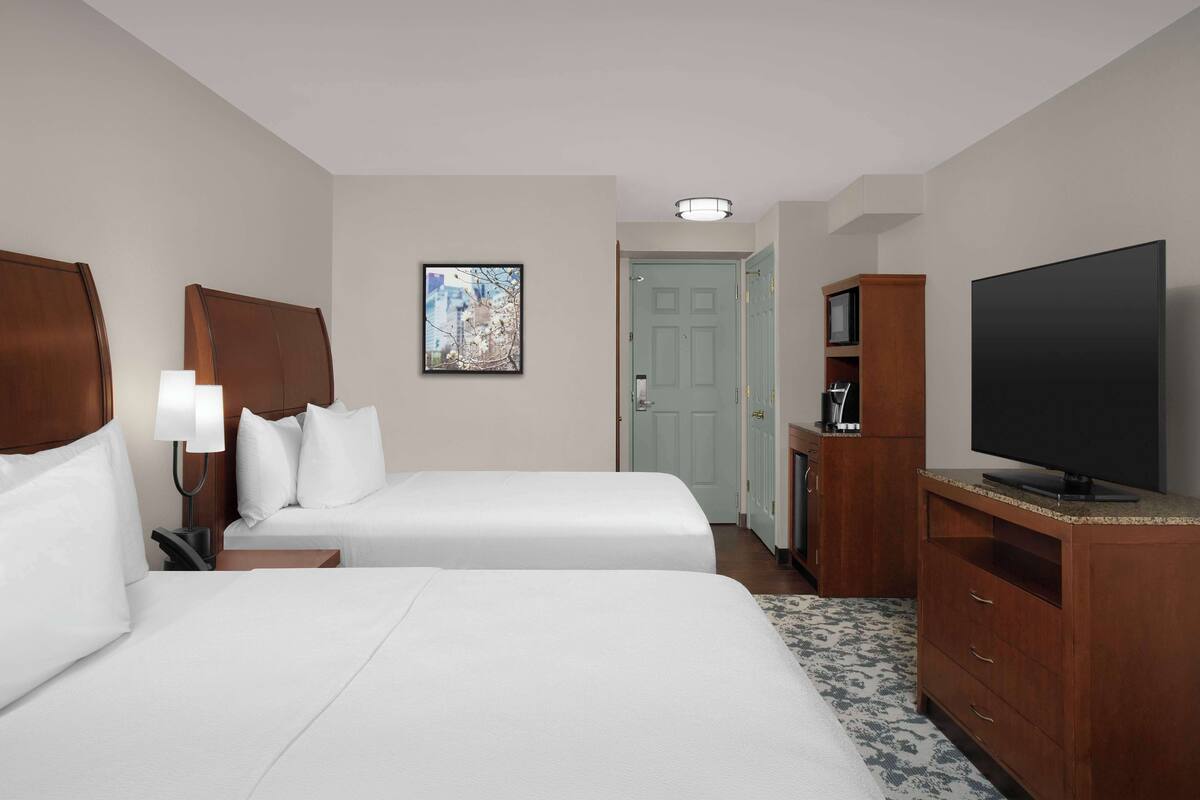 A hotel room with two beds, a television, a desk with a lamp, a framed picture on the wall, and a door leading to another room.