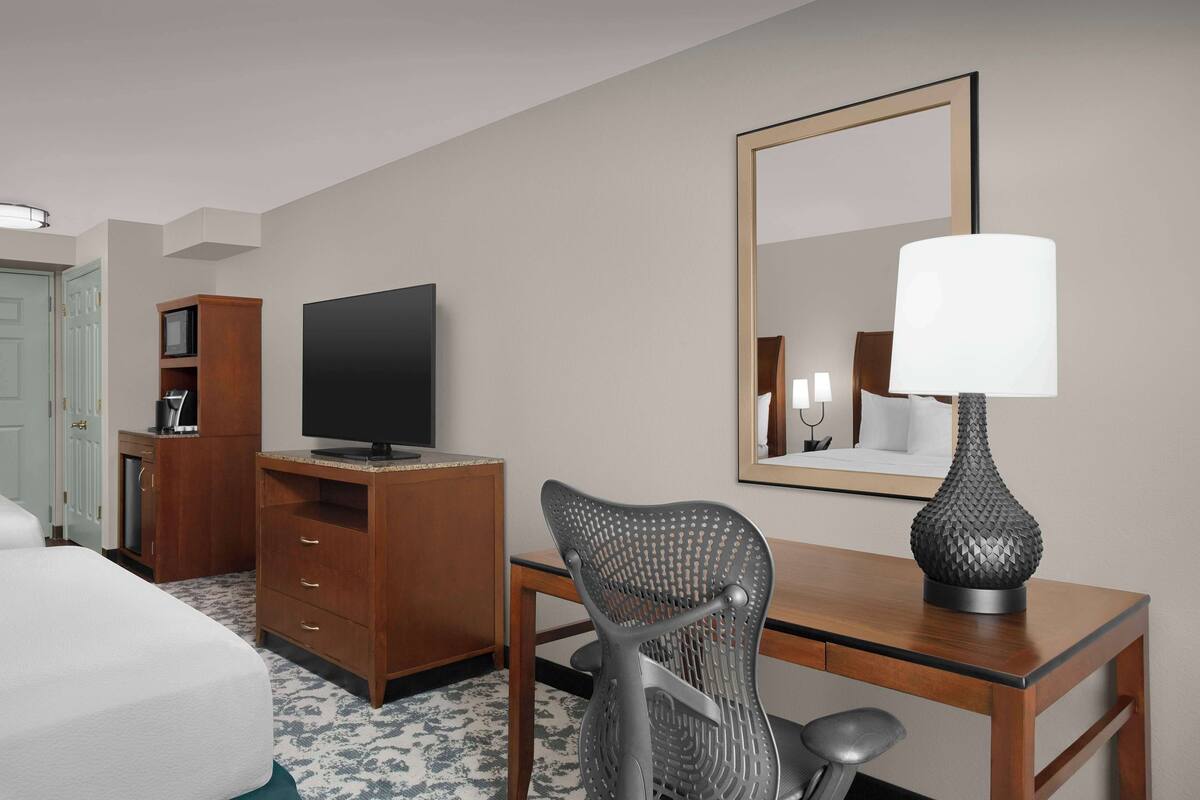 A hotel room with a bed, a desk with a lamp, a television, a chair, and a wardrobe.