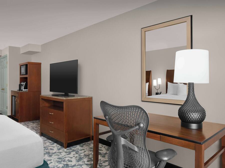 Quarto de hotel com uma cama, escrivaninha com luminária, televisão, cadeira e guarda-roupa.